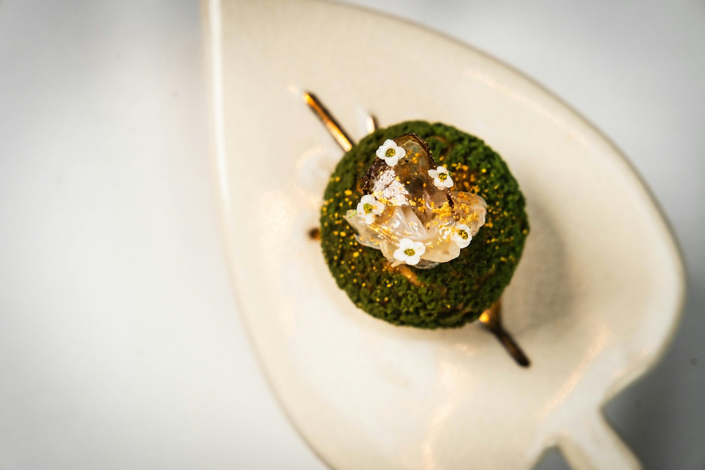 Pasticcino verde sferico con fiori eduli e foglia d'oro su un piatto a forma di foglia.